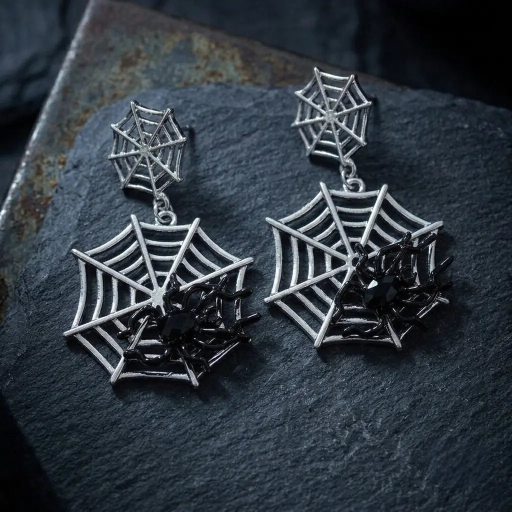 Pair of gothic silver-tone double spider web drop stud earrings resting on dark grey rough slate rock with irregular layered edges — small web stud and large web pendant with black enamel spider — dark blue-grey stone background with soft natural side lighting

