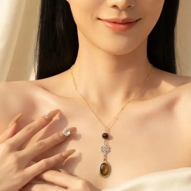 Smiling Asian woman wearing gold Y-drop cable chain necklace with tiger eye bead, rhinestone floral connector and oval tiger eye cabochon pendant, hand resting on chest, white strapless top, warm champagne silk drape background with soft sunlight

