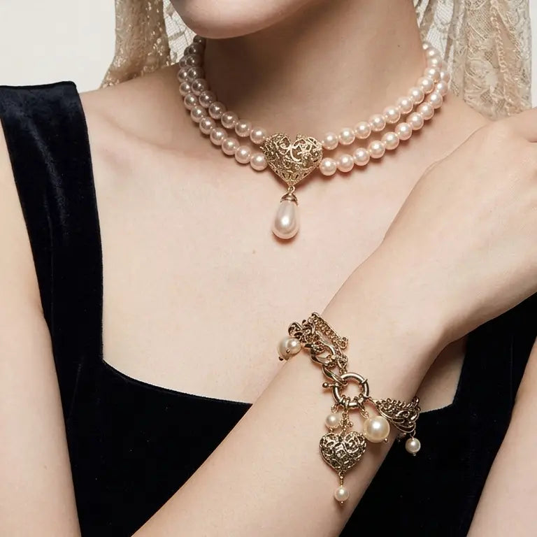 Woman with light skin tone and gold lace hair accessory wearing vintage baroque rose gold multi-chain pearl heart charm bracelet on wrist, paired with matching double-strand pearl necklace with baroque heart pendant and teardrop pearl drop at neck, black velvet strapless top, warm beige background, AI-generated model image — bracelet shown as part of coordinated set

