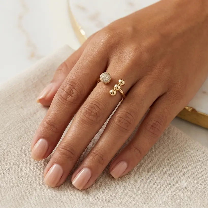 Woman wearing gold alloy open band ring with micro-pave zirconia ball and three gold bead ends on index finger, hand resting on cream linen fabric with white marble and gold book spine in background

