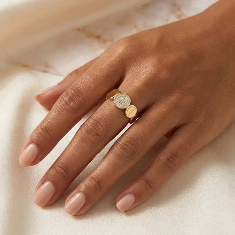 Woman wearing gold alloy wide band ring with alternating micro-pave zirconia and smooth gold oval discs on middle finger, hand resting on cream satin fabric with marble background

